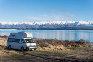 Image of a campervan parked beside a lake with a beautiful view of a mountain range.  