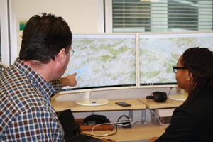 A geospatial expert walks a graduate through some of their work, pointing to some mapping data on screen