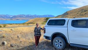 A woman standing beside a ute, with mountains and a lake in the background