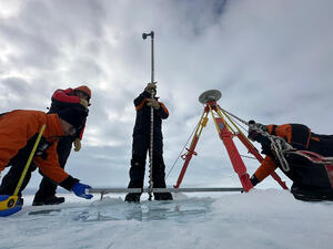 A team of four people with tools surrounding a hole in the ice, working on a tide guage