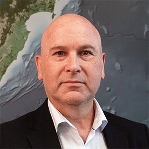 Headsot of Andrew Pilcher wearing a white shirt and dark jacket in front of a map of New Zealand