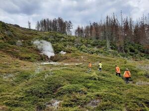 Four people in high-vis vests walk towards a vent of steam coming out of a hillside