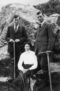 Two men dressed in suits stand behind a seated woman. Both men are holding pickaxes and there is a mountain behind them.