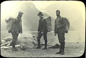 Three men standing in front of a river and mountains. Each man is wearing a large backpack.