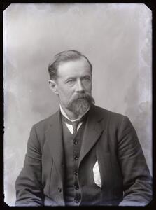 A portrait of a man with a beard and moustache. He wears a suit.