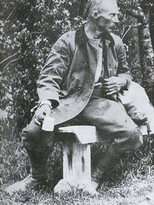 An older man sitting on a bench with native bush behind him. He is holding a tin cup and appears to be leaning to talk to someone to his left.