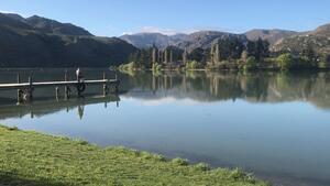 View of a peaceful lake from the shore. A man is fishing from a jetty.