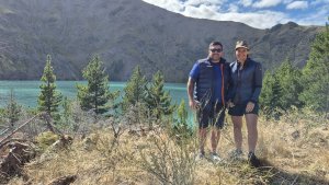 Two people stand in front of a lake. Wilding pines can be seen behind them, and the branches of a whip broom can be seen in front of them