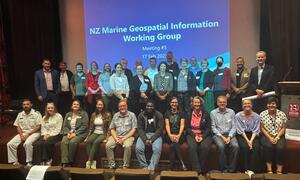A large group of people standing in front of a screen saying NZ Marine Geospatial Information Working Group meeting 5