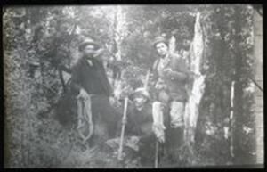 Three men posing in amongst NZ native bush. Each one holds mountaineering gear such as a coil of rope or a pick. The photo is low quality from age.