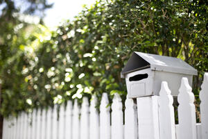 A letterbox sitting on a picket fence with a sunny garden behind it.
