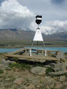 A black and white trig beacon on a hill looking across a lake and mountain range.